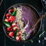 Blueberry smoothie topped with strawberries, pink blueberries, popped quinoa, on a black tile background surrounded by strawberries and blueberries.