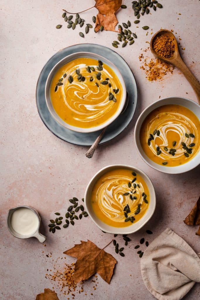 Three bowls of butternut squash soup with pumpkin seeds and fall leaves.