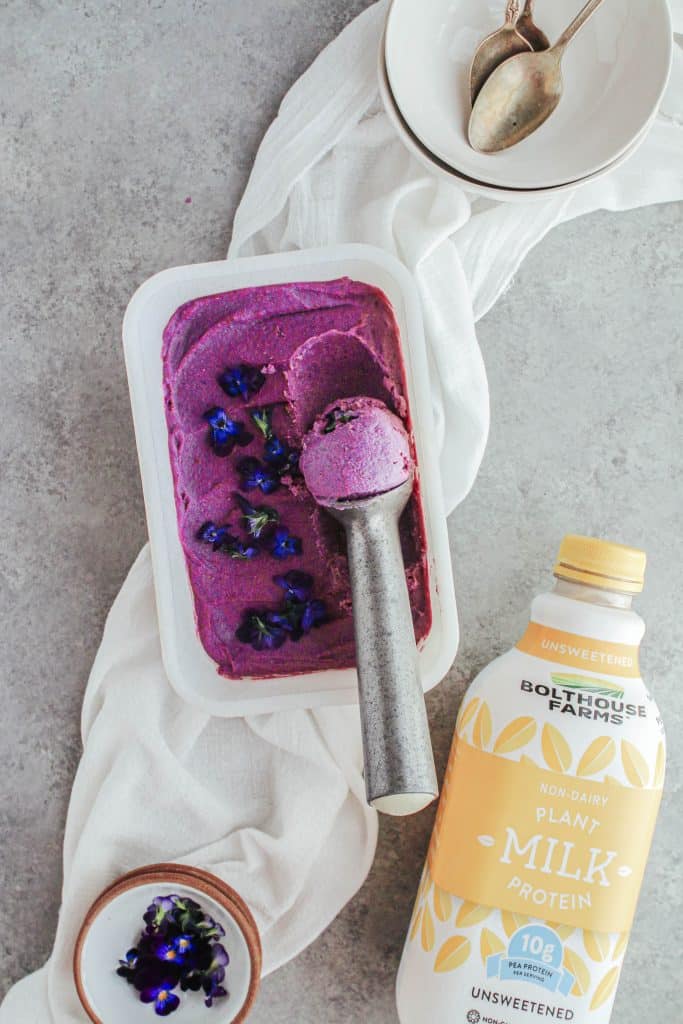 Purple nice cream with an ice cream scoop a bowl of edible purple flowers and a bottle of plant milk.