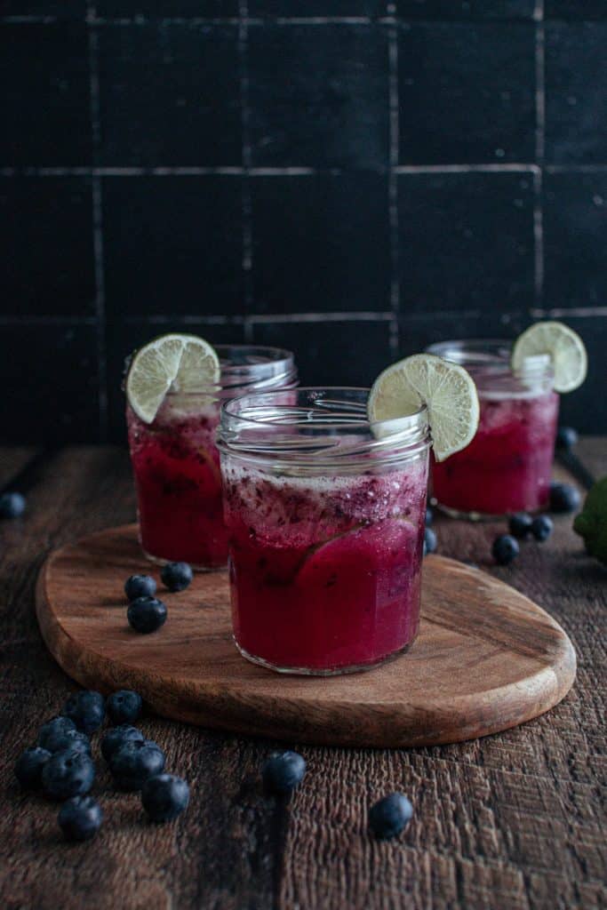 Three glasses of blueberry limeade on a wooden cutting board.