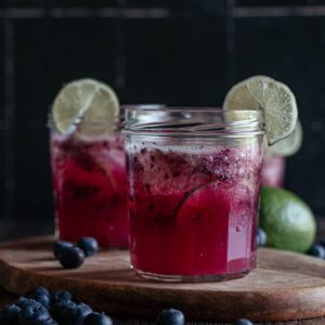 Two glasses of blueberry limeade on a wooden cutting board.