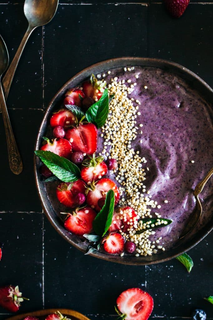 Blueberry smoothie bowl topped with strawberries, pink blueberries, popped quinoa, on a black tile background surrounded by strawberries and blueberries.