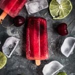 Cherry limeade popsicles on a gray surface with lime wedges.