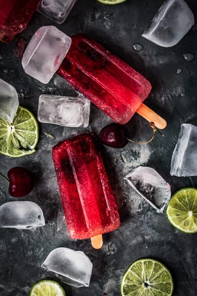 Two cherry limeade popsicles with fresh cherries and limes.