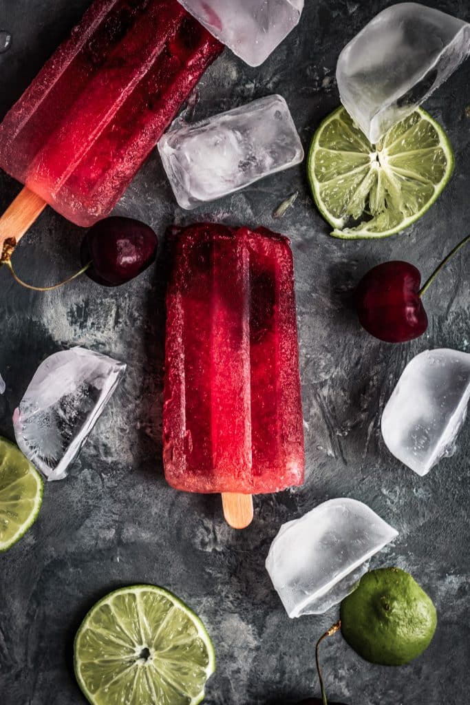 Cherry limeade popsicles on a gray surface with lime wedges.