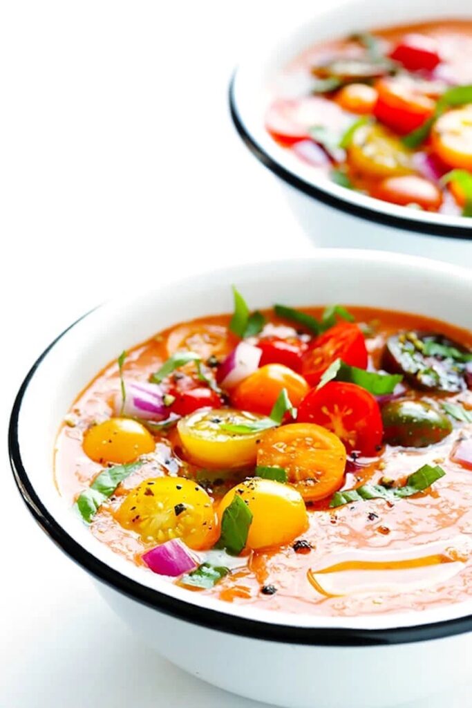 Close up of black rimmed bowl with deep orange gazpacho topped with sliced heirloom tomatoes.