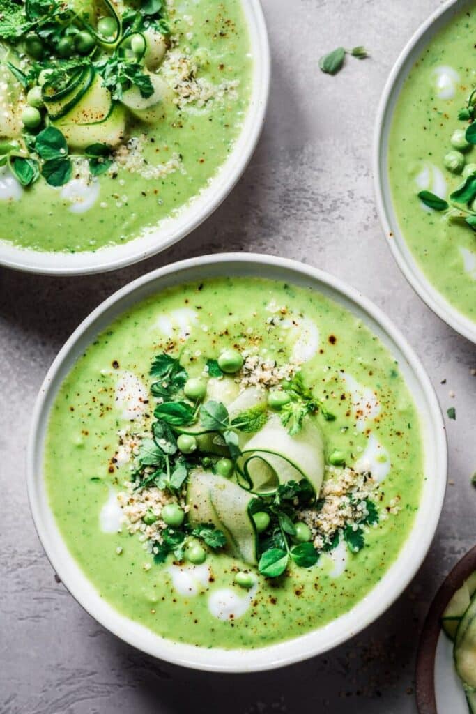 Three bowls filled with light green cold soup topped with peas, shaved cucumber, and coconut cream.