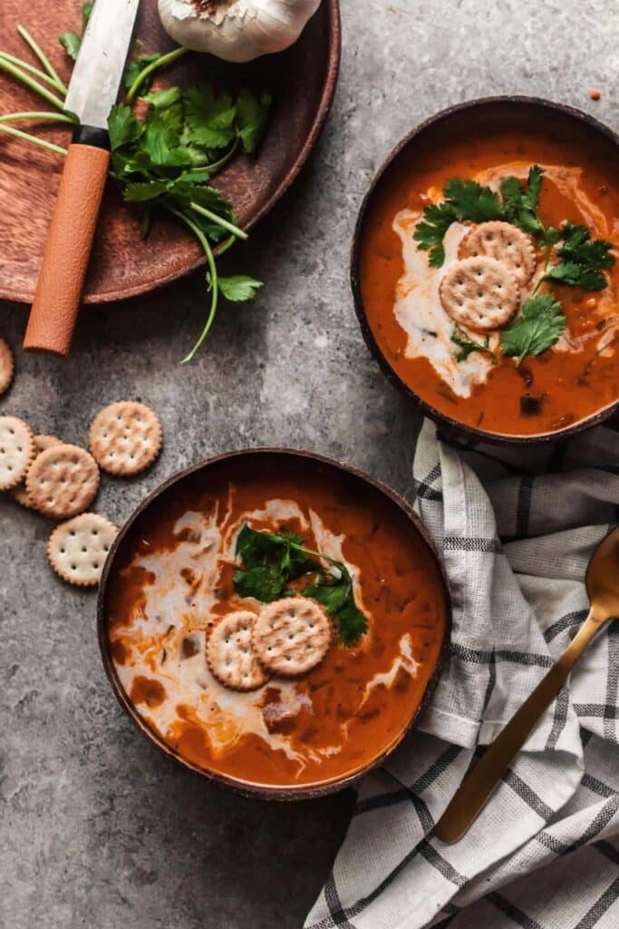 Two coconut bowls filled with red tomato lentil soup with crackers on top.