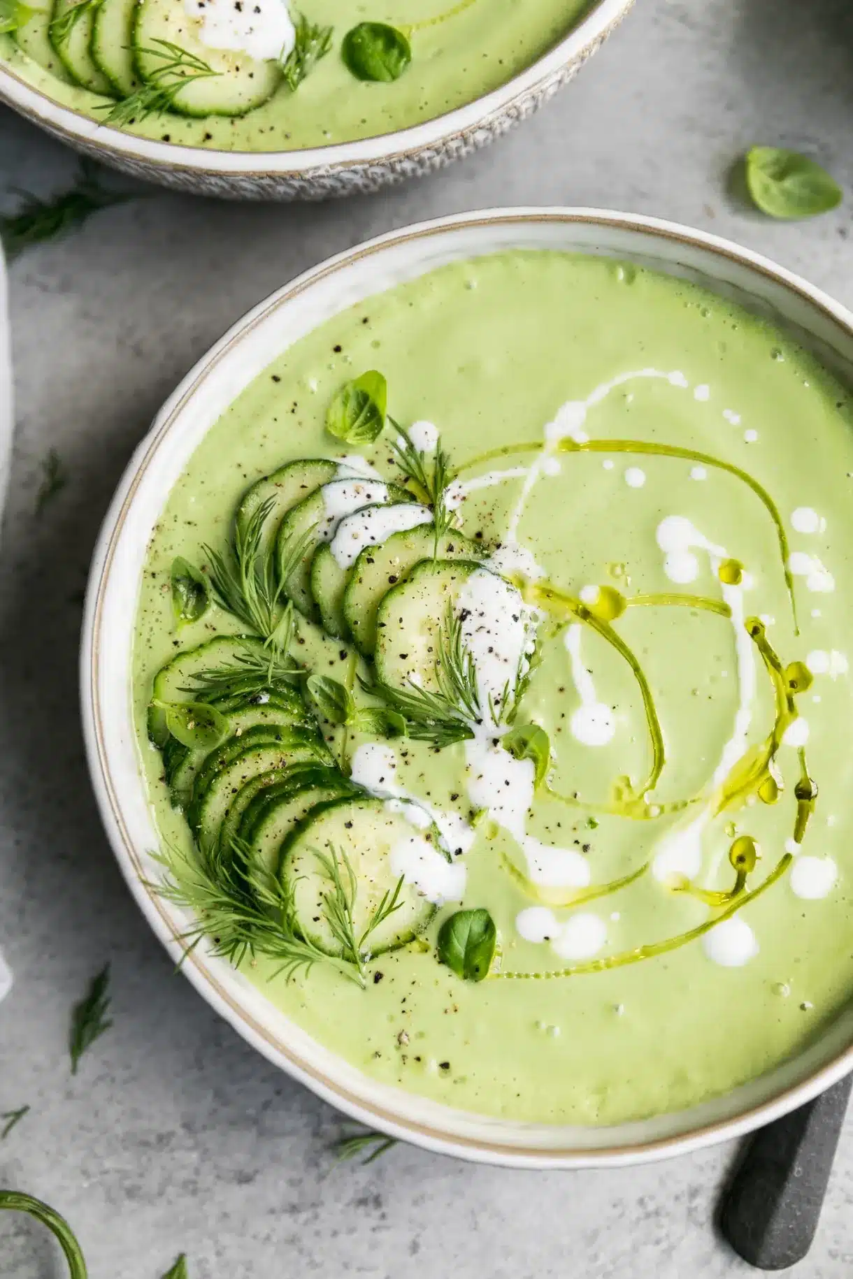 Large bowl of light green creamy soup with olive oil drizzle and fresh cucumbers on top.