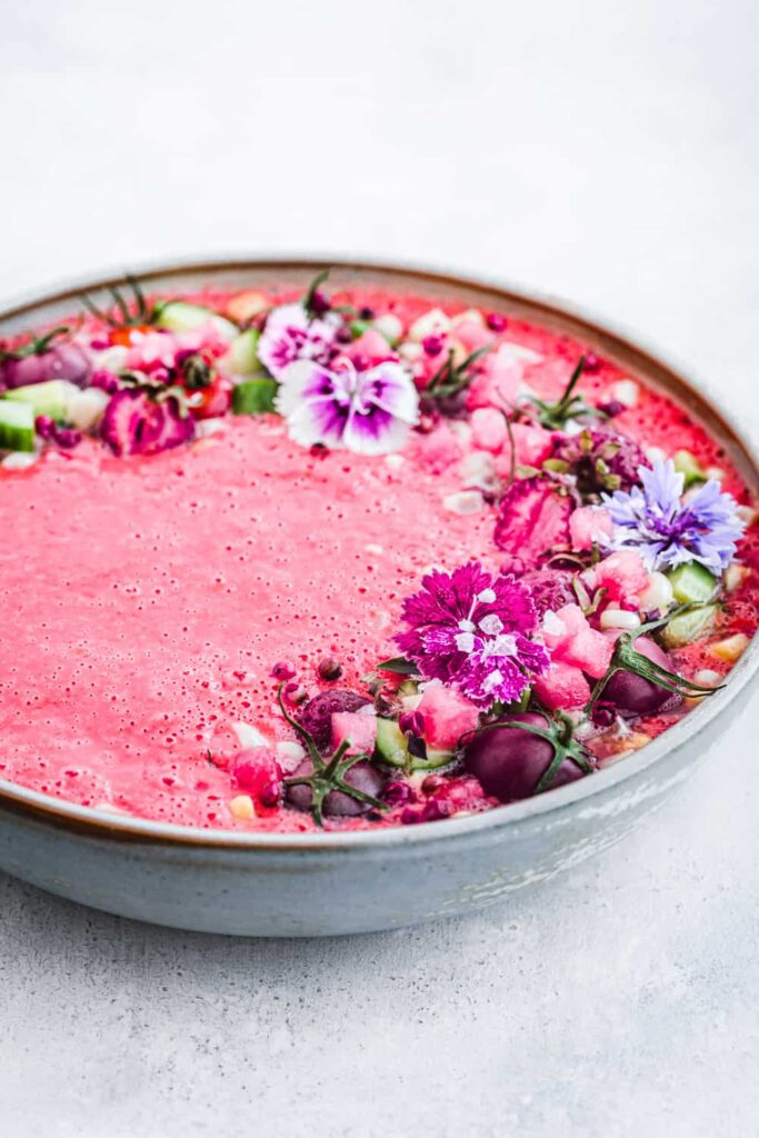 Large bowl of bright pink watermelon gazpacho topped with edible flowers. 