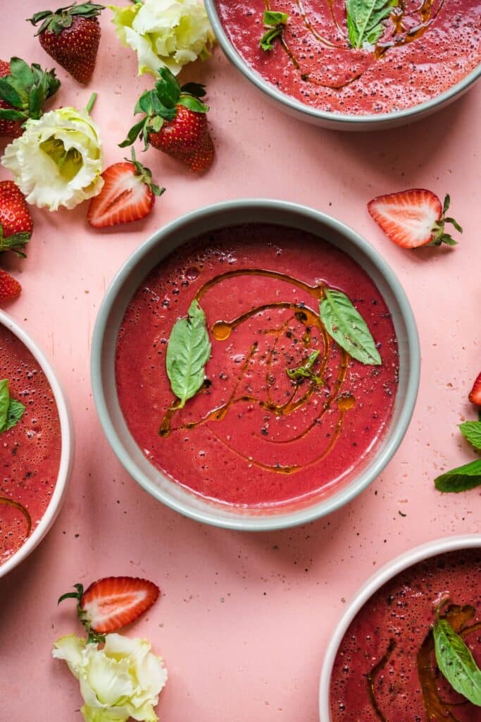 Bowls of deep pink-red gazpacho with olive oil drizzle and fresh basil on a pink surface. 