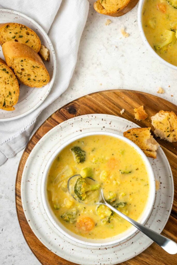 Large bowl of cheesy broccoli soup on a round wooden board with bread on it.