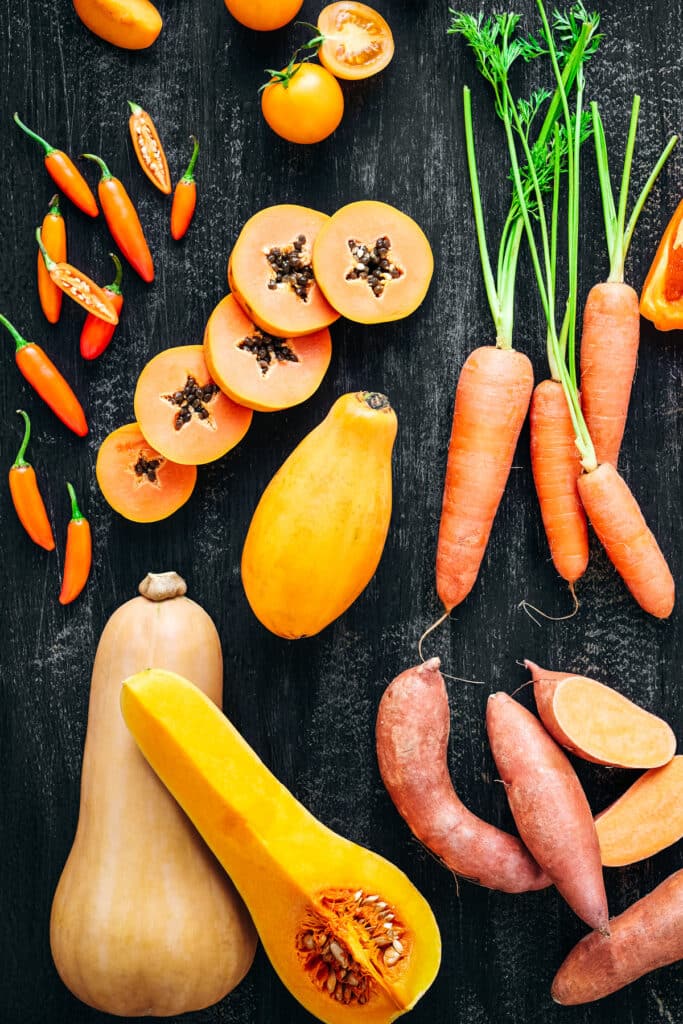 Butternut squash, carrots, persimmon, sweet potato, and orange peppers on a black backdrop.
