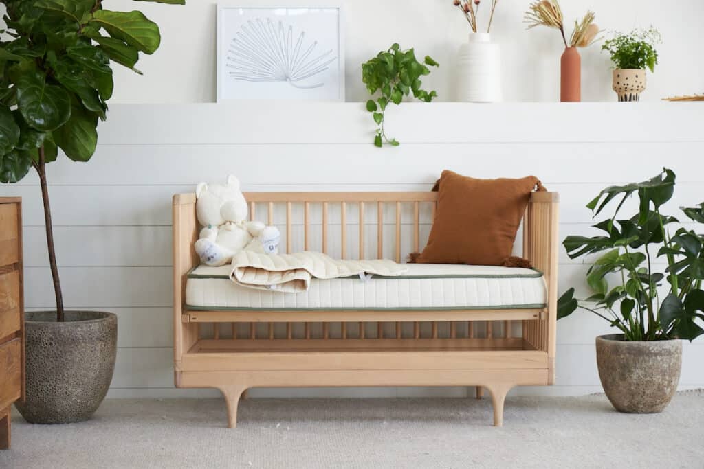 Crib mattress in modern crib with cream blanket and rust colored pillow.