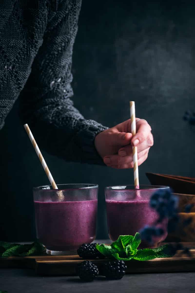Two cups of blackberry smoothie with a woman stirring one of the glasses with a paper straw.