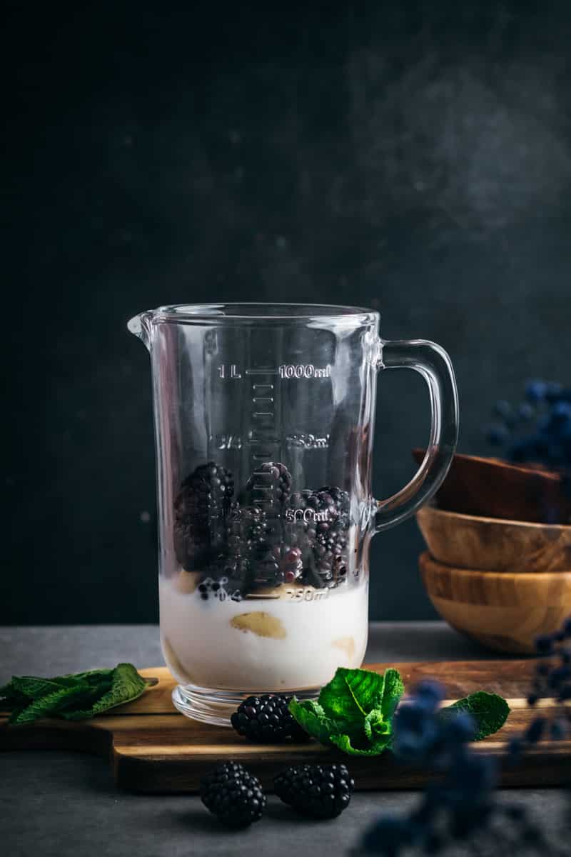 Ingredients for blackberry smoothie in a blender with fresh blackberries and mint on the side.