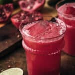 Two clear glass cups filled with pomegranate limeade surrounded by limes and pomegranate arils.