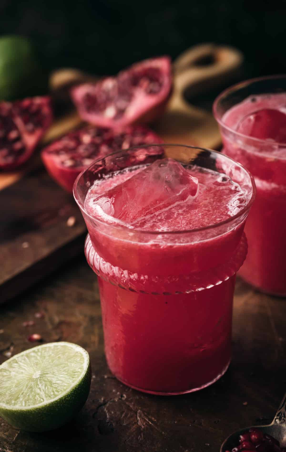 Two clear glass cups filled with pomegranate limeade surrounded by limes and pomegranate arils. 