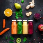 Bright orange, green, yellow, and dark pink vegetable smoothies with fresh fruit and veggies around.