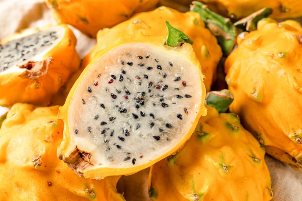 Pile of yellow dragon fruit with one sliced in half showing the translucent white flesh and small black seeds.