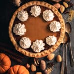 Pumpkin pie with seven swirls of whipped cream surrounded by small pumpkins and spices.