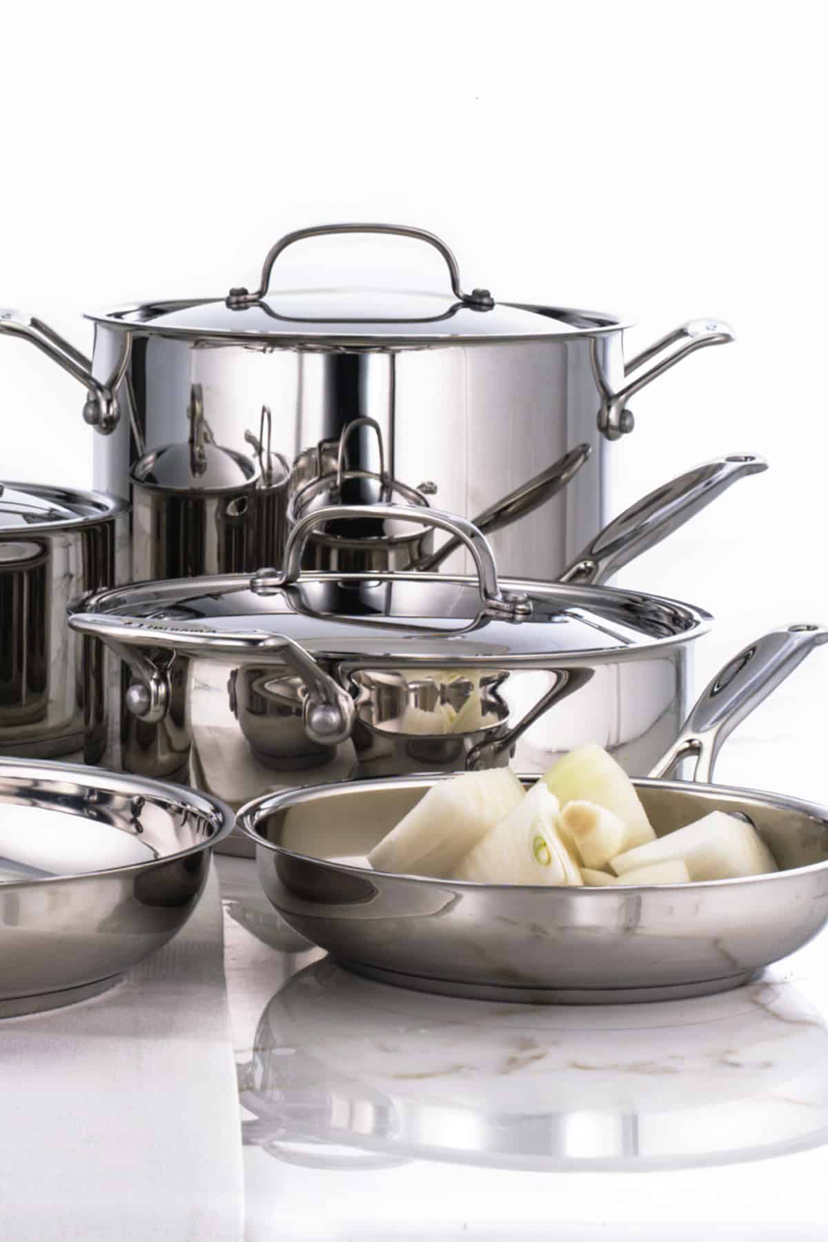 Stainless steel frying pan with white onions in it and other stainless steel pots behind it. 