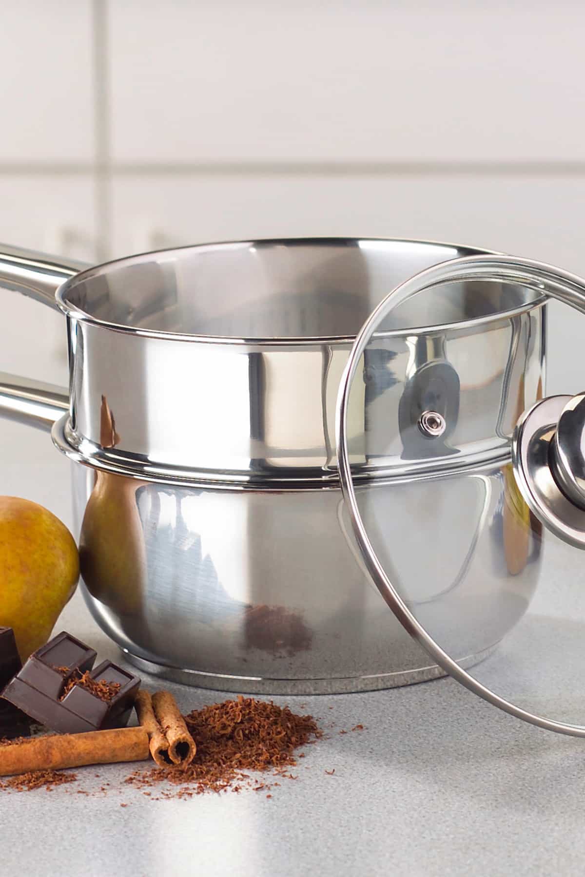 Stainless steel double boiler with a glass lid and chocolate, cinnamon and a pear next to it. 