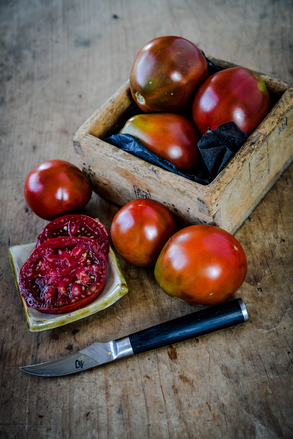 Black tomatoes in a wooden box with a few on the table. One is cut into slices in showing the inside flesh and seeds.