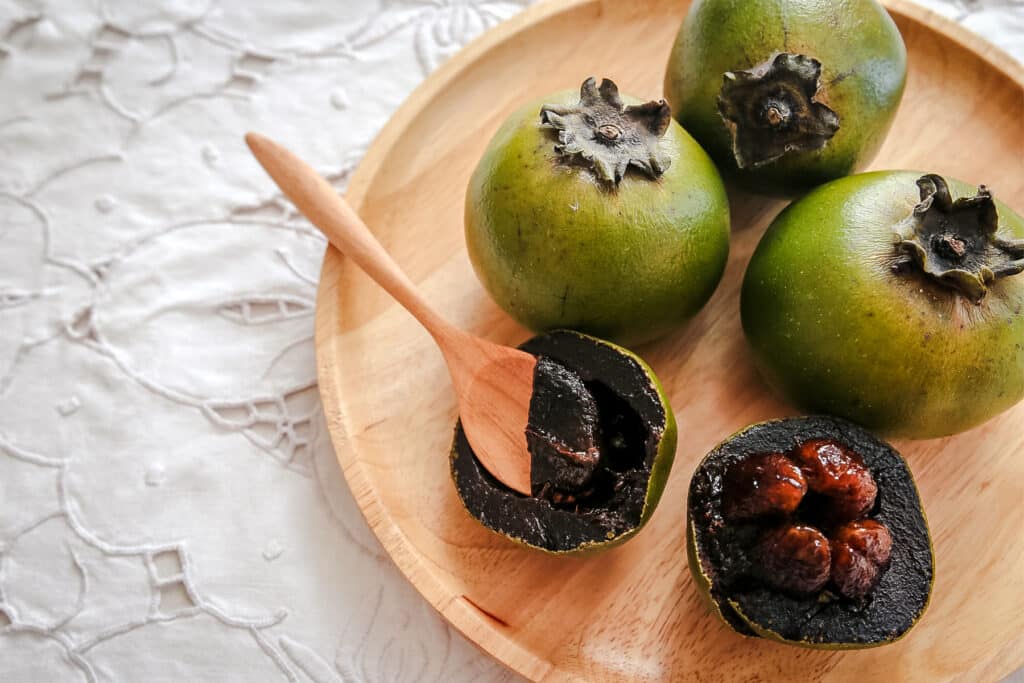 Four black sapote with one cut in half on a wooden plate with a wooden spoon.