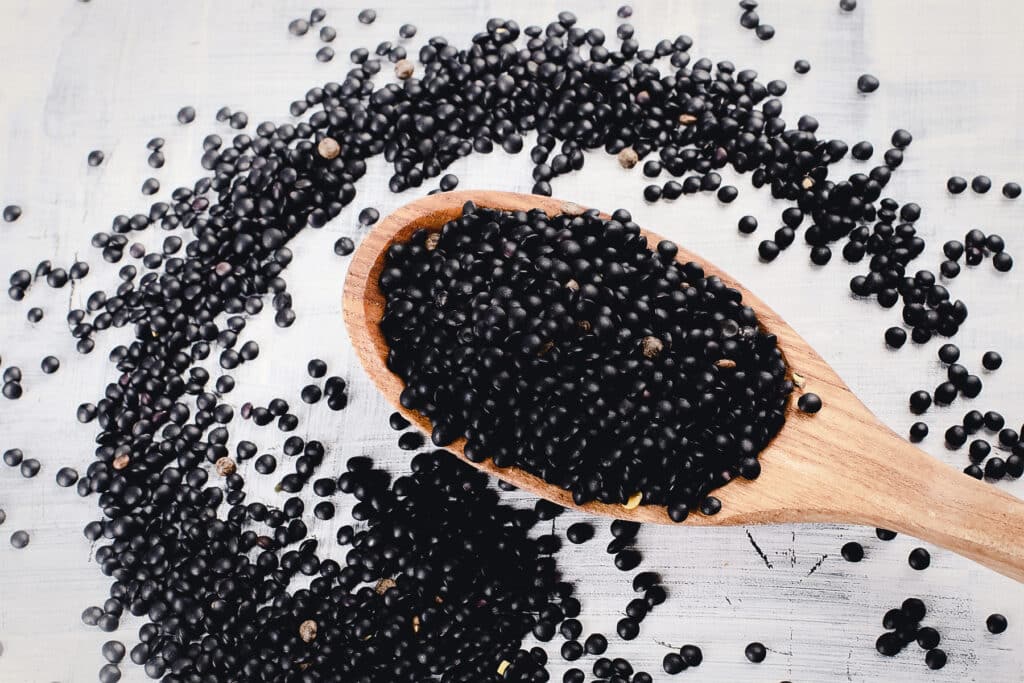Black disk shaped lentils in a wooden spoon spilling over onto the white tabletop.