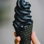 Person holding deep black soft serve ice cream in a black waffle cone.