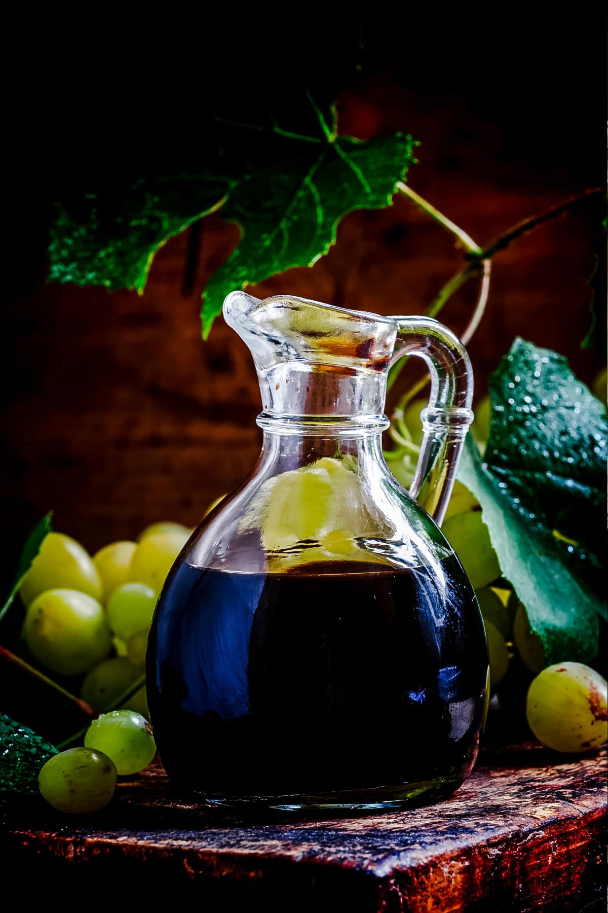 Dark balsamic vinegar in a small glass pitcher with grapes on the table next to it.
