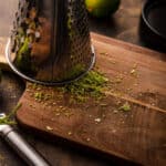 Metal box grater on top of a wooden cutting board with lime zest scattered across it.