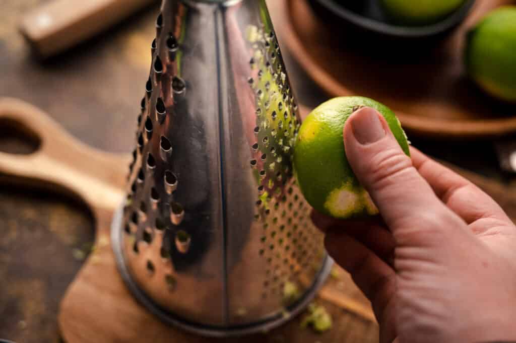 Woman holding a lime and zesting it using a circular metal box grater.