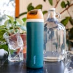 Owala teal water bottle with an orange lid on a black tile surface next to a water jug and glasses.