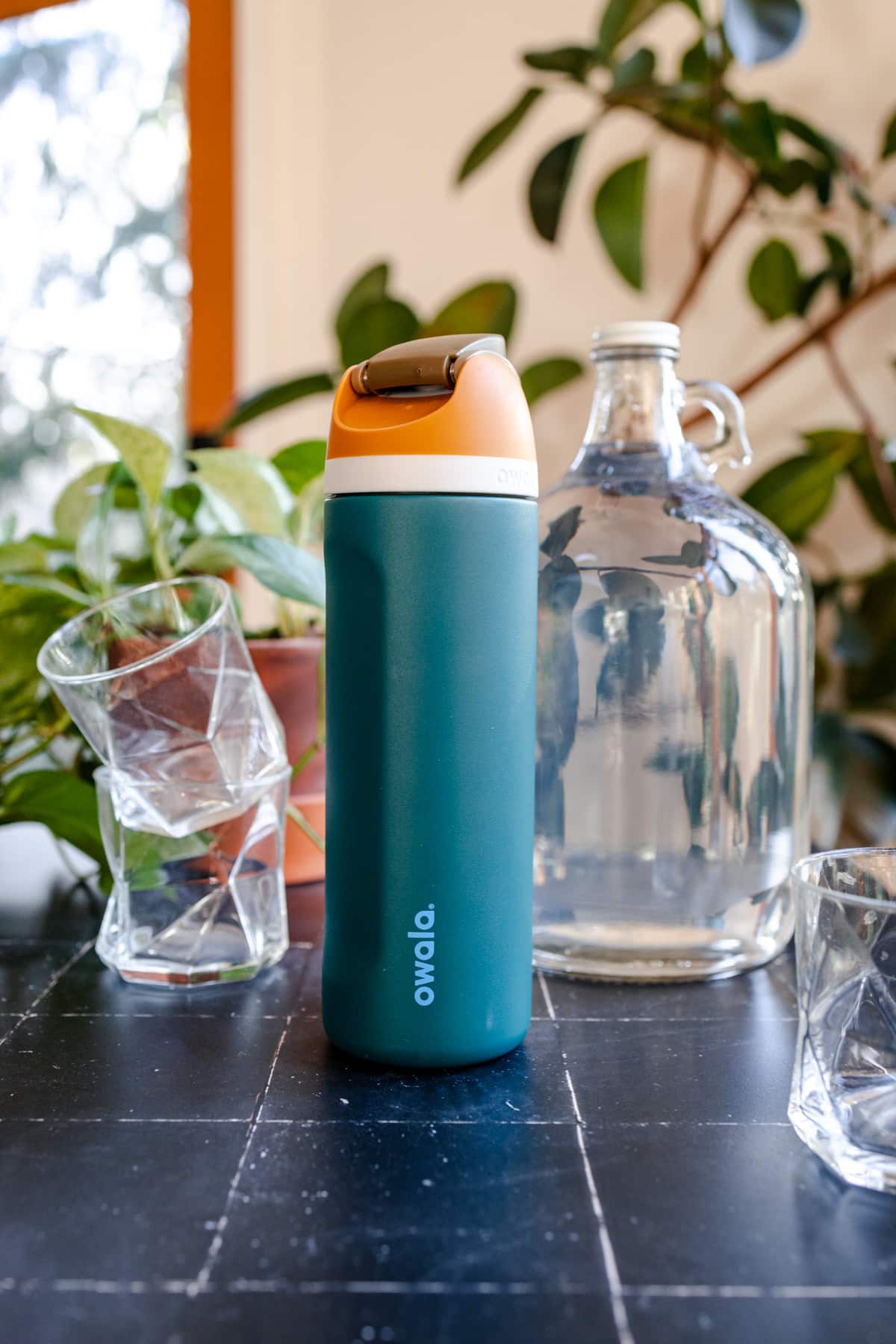 Owala teal water bottle with an orange lid on a black tile surface next to a water jug and glasses.