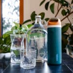 Glass gallon size jug filled with water next to three glass cups and an insulated water bottle.