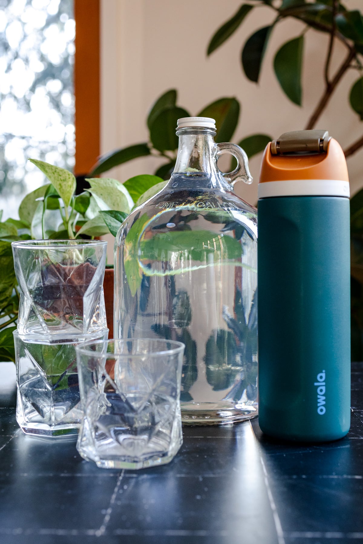 Glass jug filled with 128 ounces of water on a black tile surface next to a teal water bottle.