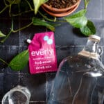 Glass gallon just and pink pomegranate Everly hydration packet on a black tile surface.