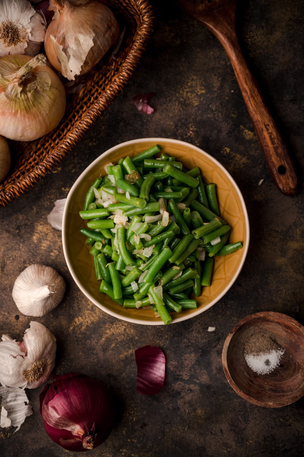 Straight down image of cooked green beans in a ceramic bowl with onions, garlic, and spices next to it.