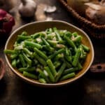 Cooked canned green beans in a ceramic bowl with fresh ingredients surrounding it.