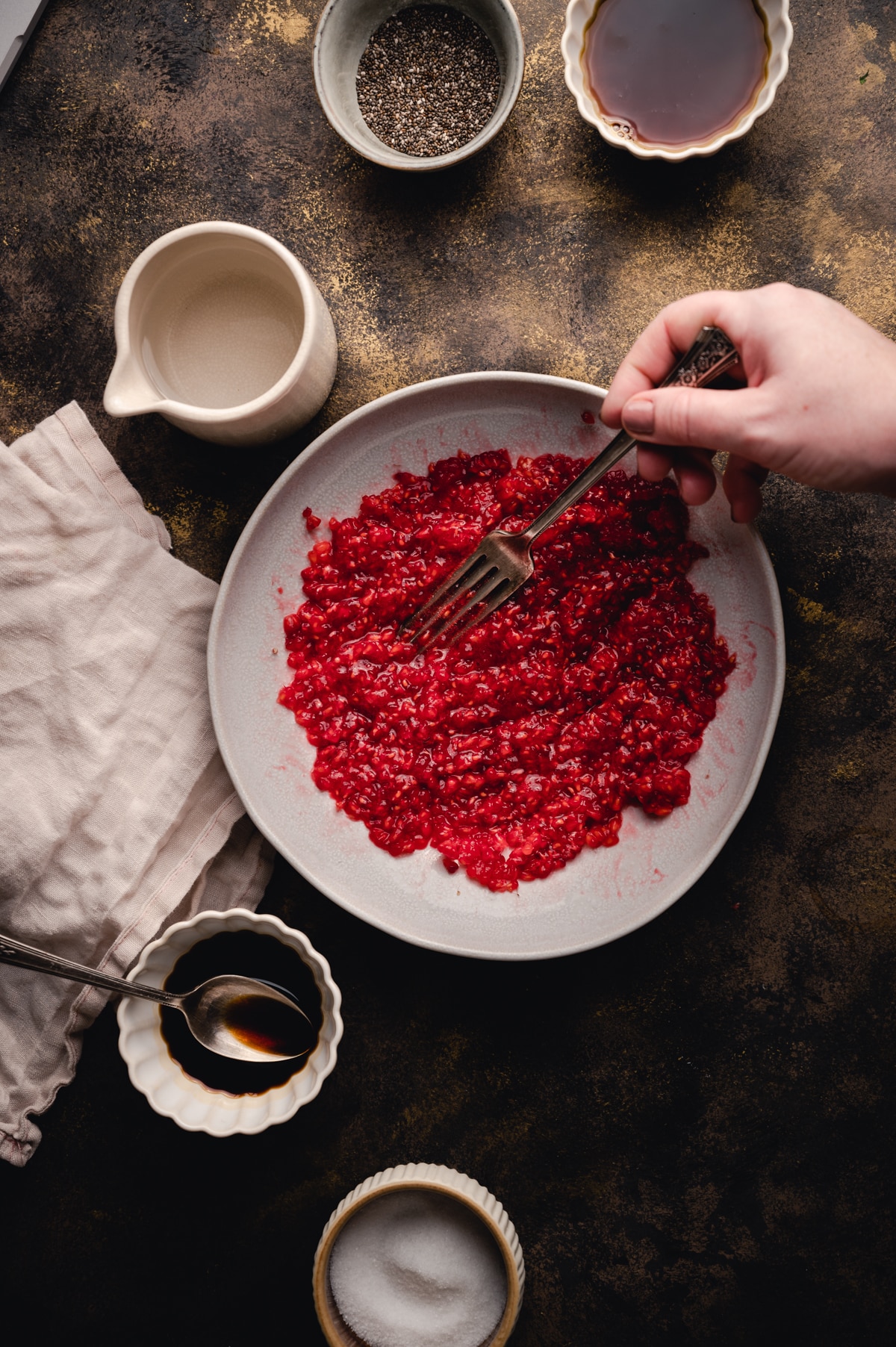 Fresh raspberries being smashed on a white plate with ingredietns in bowls surrounding it.