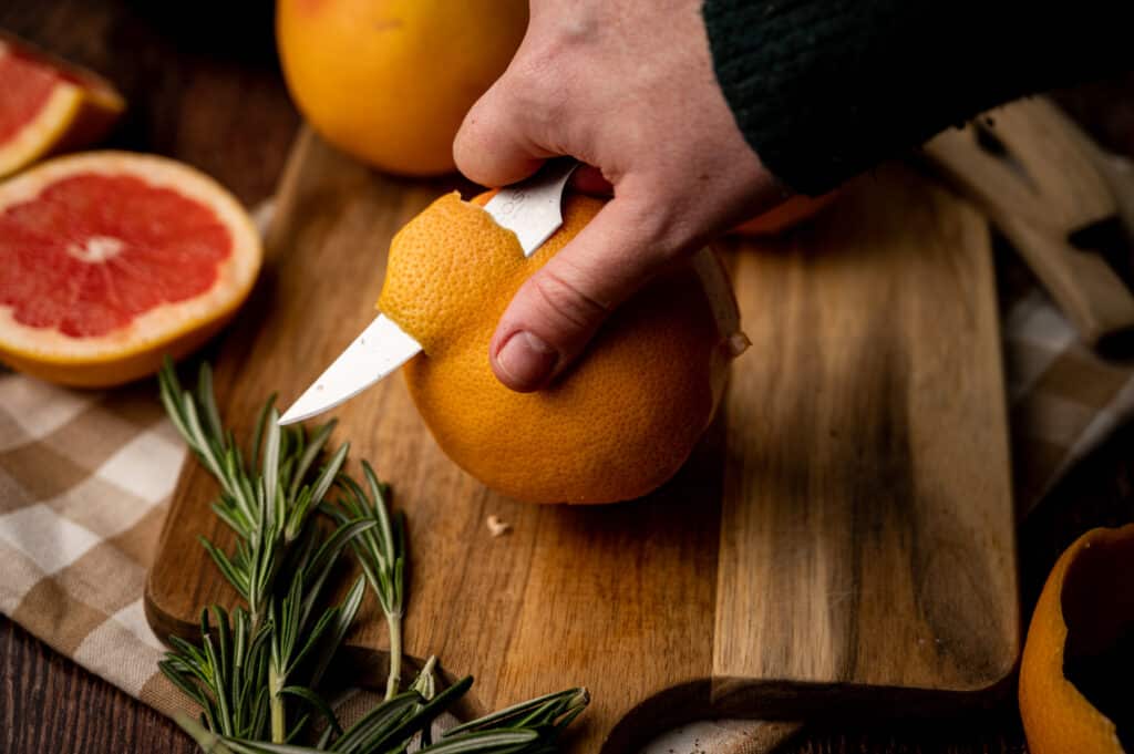 Peeling a grapefruit with a paring knife on a wooden cutting board.