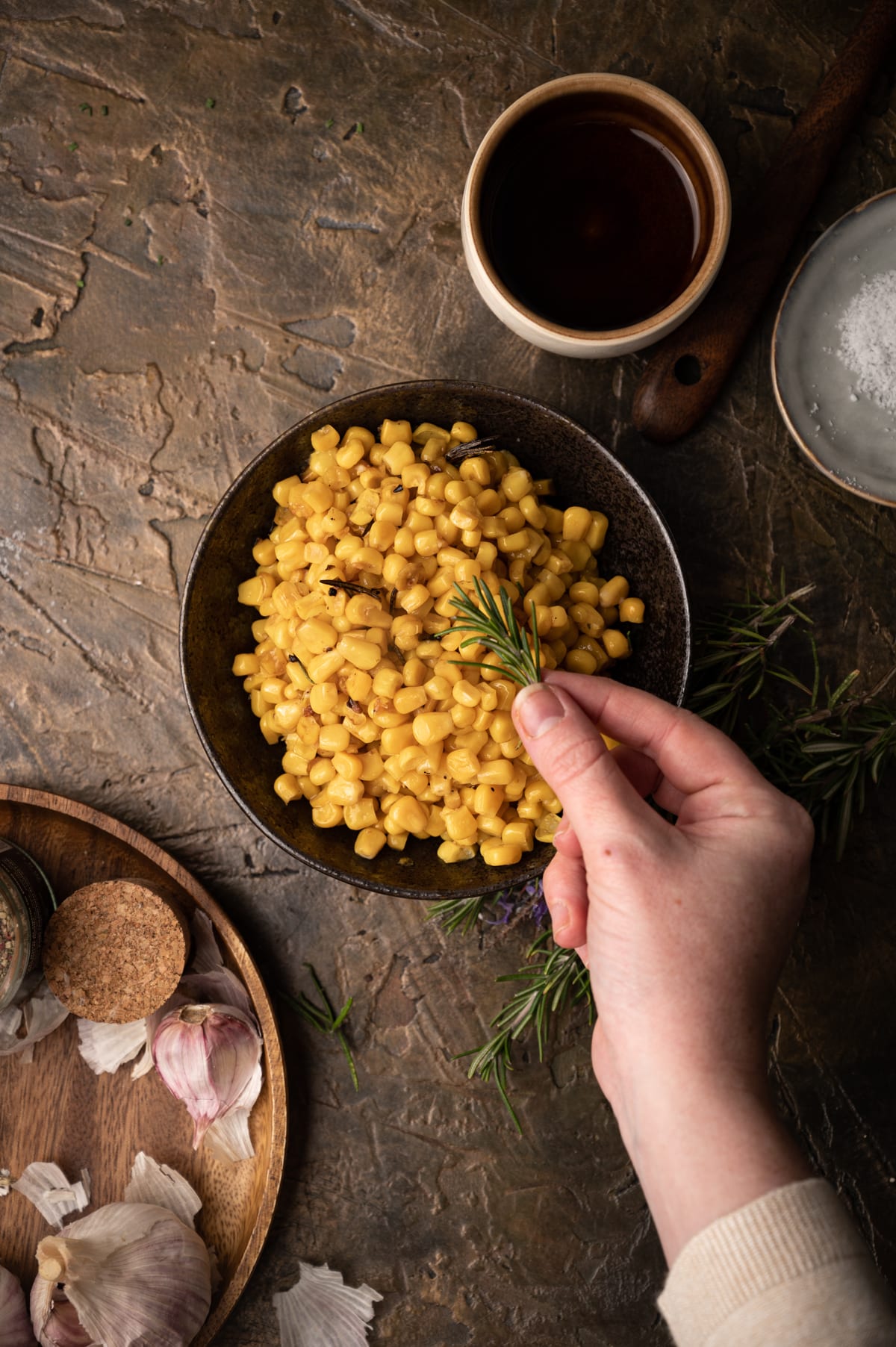 Bronze bowl of freshly cooked canned corn with a woman placing fresh rosemary on top.