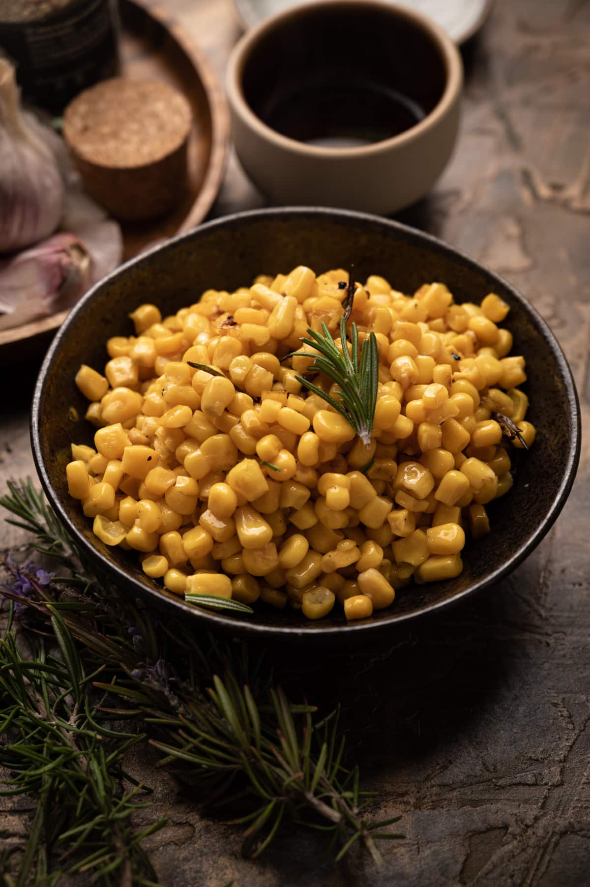 Freshly cooked corn in a bronze ceramic bowl with fresh rosemary sprigs laying nearby.