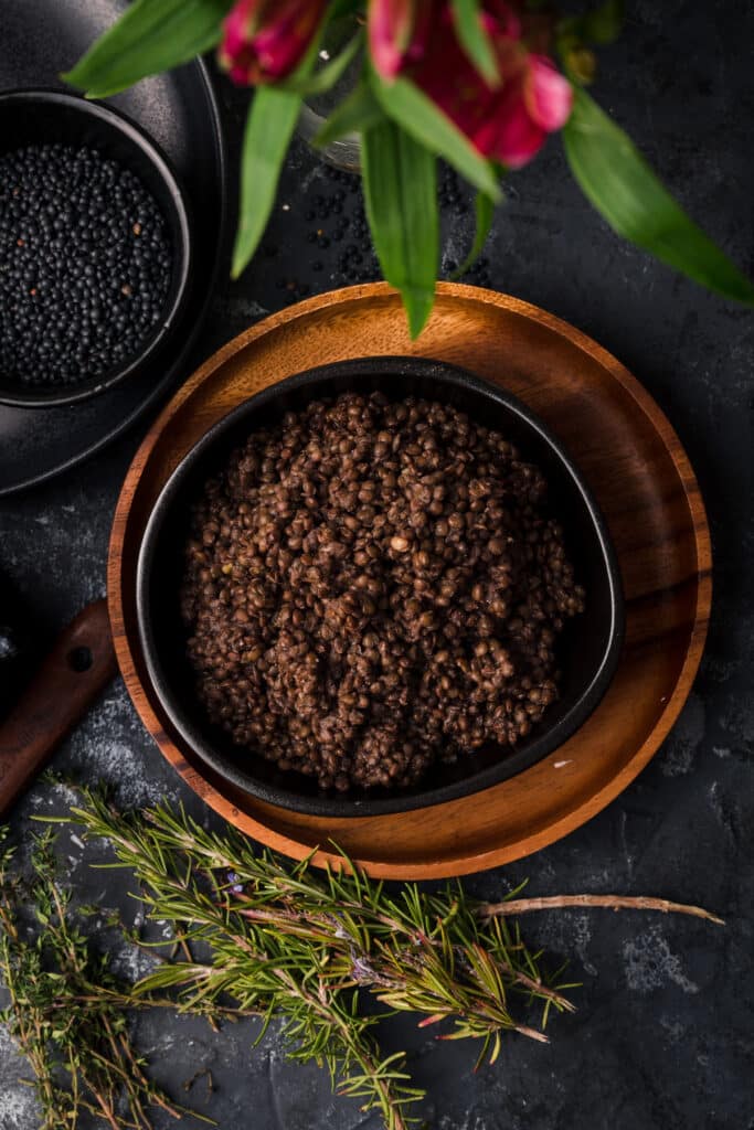 Black ceramic bowl filled with freshly cooked lentils on a wooden plate with fresh herbs scattered around.