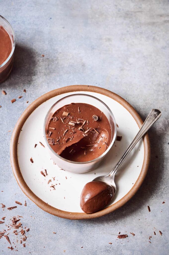 Creamy vegan chocolate mousse in a clear glass bowl on a white plate with a spoon of mousse on it. 