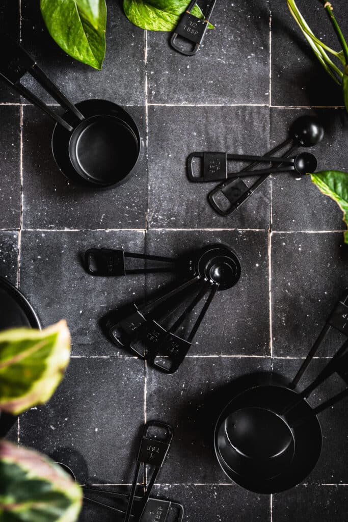 Black measuring spoons and measuring cups on black tile with silver grout.