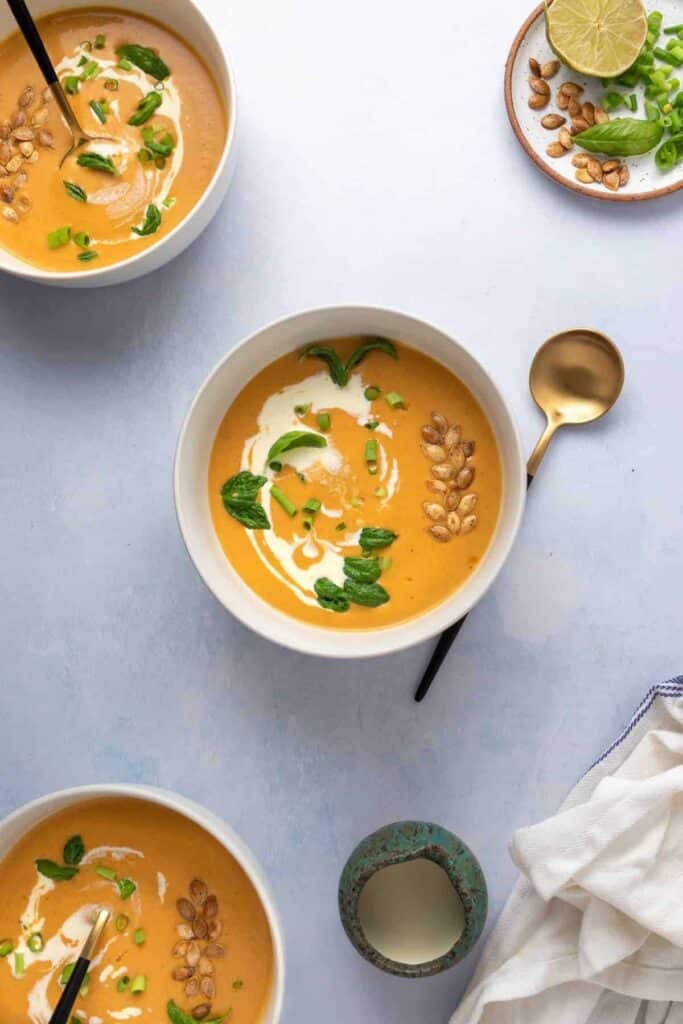 Three bowls of orange colored soup with coconut cream and fresh herbs on top.