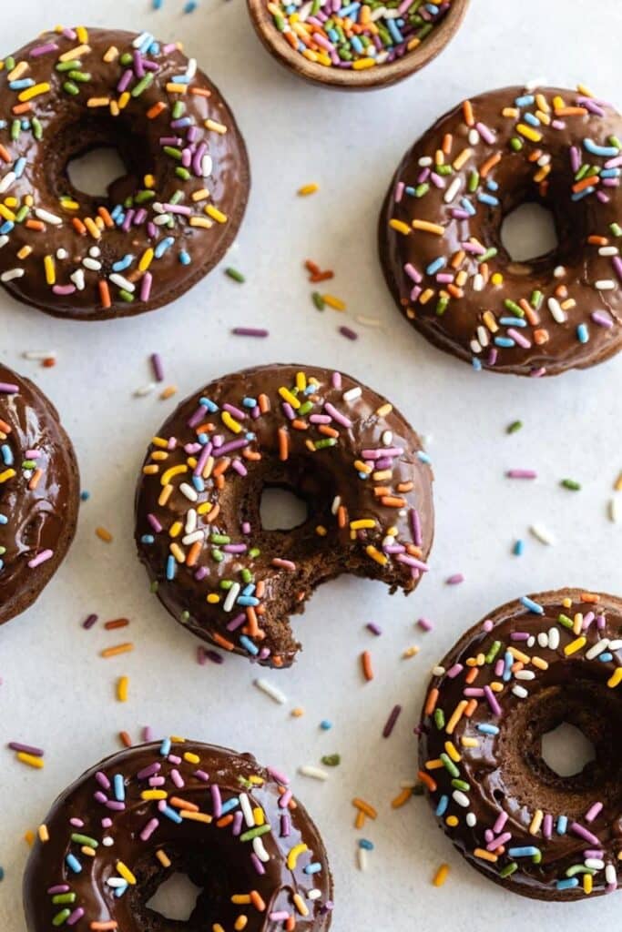Several baked chocolate donuts covered in rainbow sprinkles.